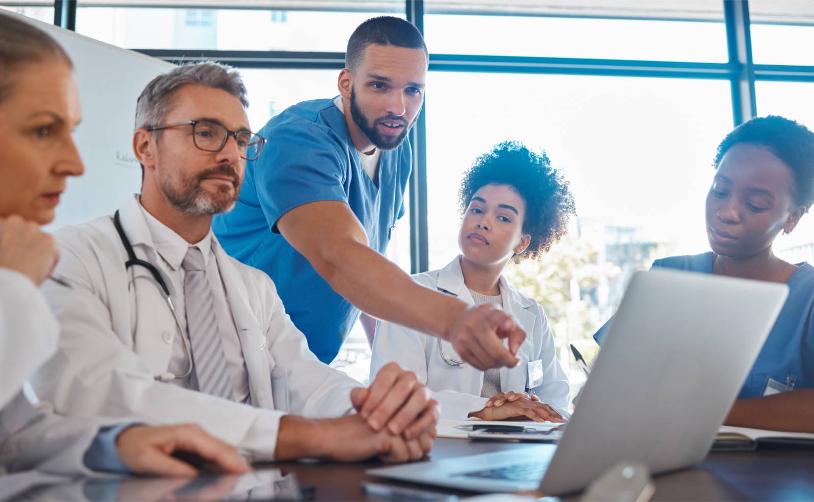 A group of doctors sitting down around conference table discussing something on a laptop screen